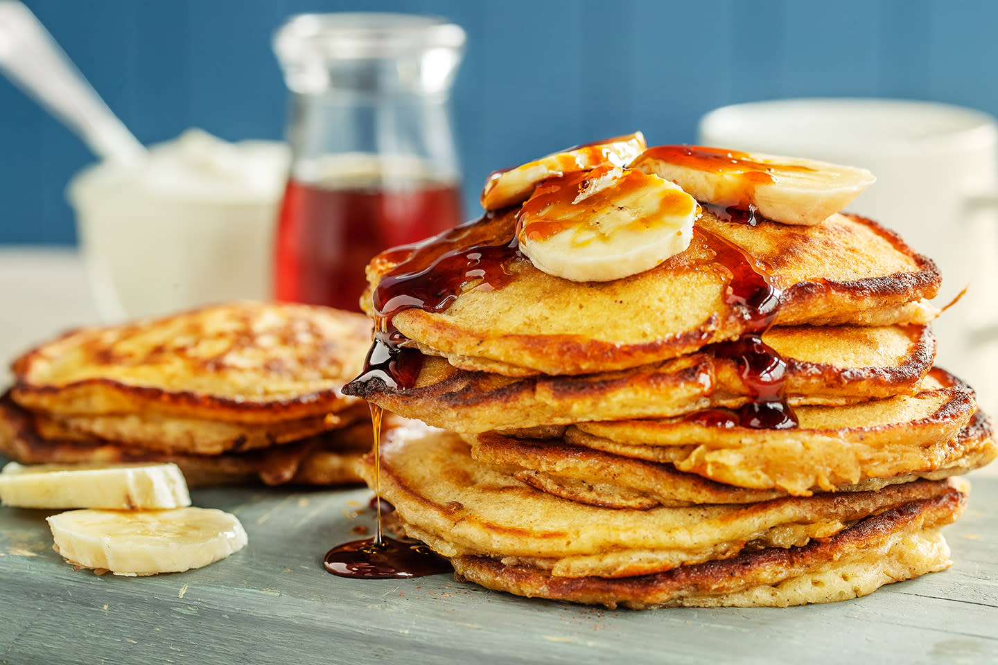 Bananpannekaker stablet på blågrønn trefjøl. Servert med skiver av fersk banan, og sirup. Flaske med sirup i bakgrunnen.