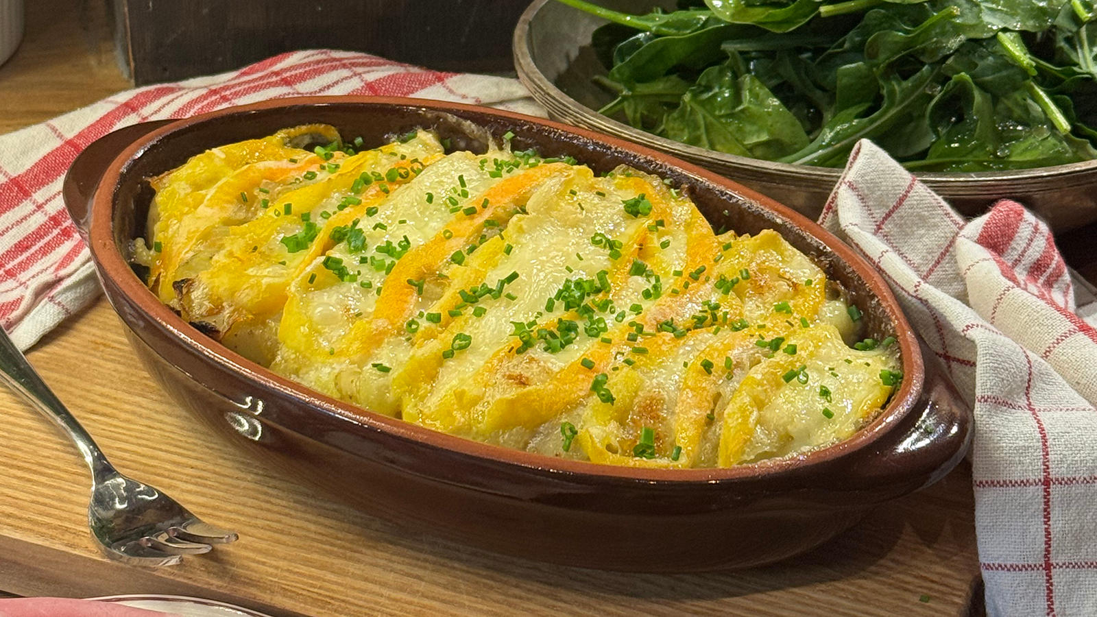 Gratinert Chaumes på knuste poteter servert i oval ildfast form på rustikk skjærefjøl med grønn salat i bakgrunnen.