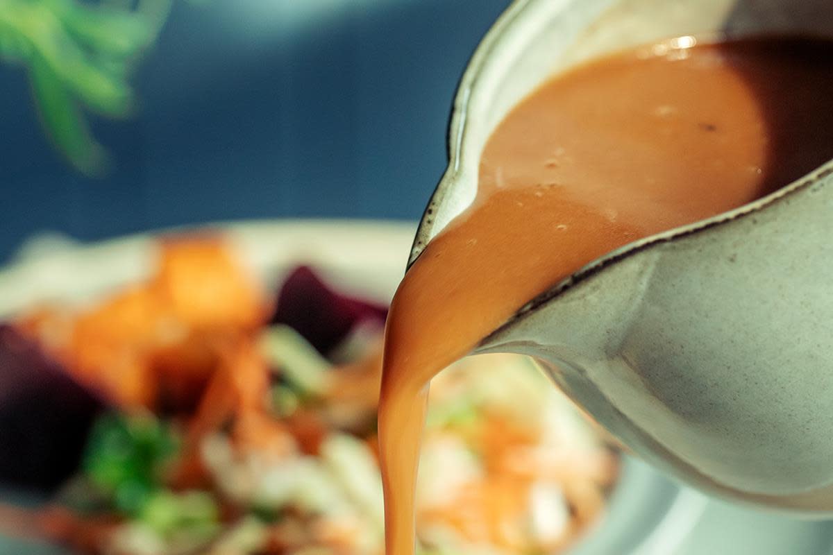 Lys brun kaffesaus helles fra lys sausemugge i stengods. Tallerken med lammemiddag i bakgrunnen.