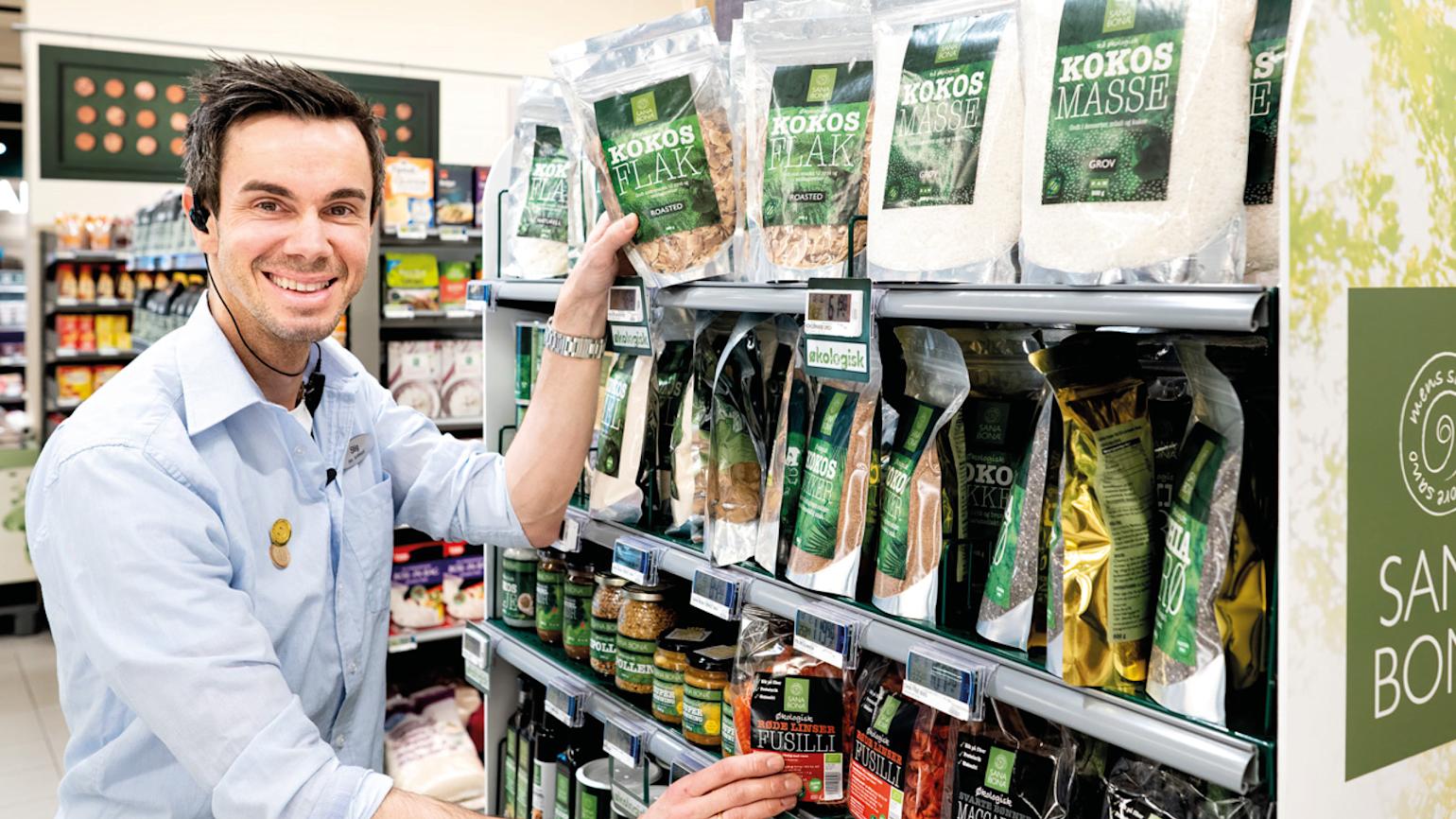 Glad fyr med lyseblå skjorte setter varer i hyllene hos en av Coop sine butikker. 