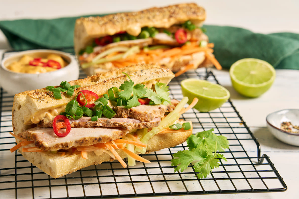 Pork belly Bánh mì med syltede grønnsaker servert i lys baguette med chiliaioli, frisk koriander og rød chili. Baguetten står på en svart rist, med en baguette til bak, og halve lime på siden.