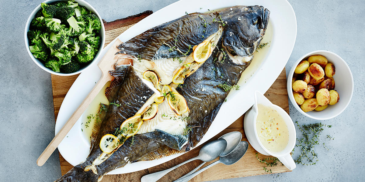 Helgrillet kveite med sitronskiver inni, servert på avlangt hvitt fiskefat på trefjøl. Skåler med brokkoli og stekte poteter ved siden av, og en sauseøse med smørsaus på siden.