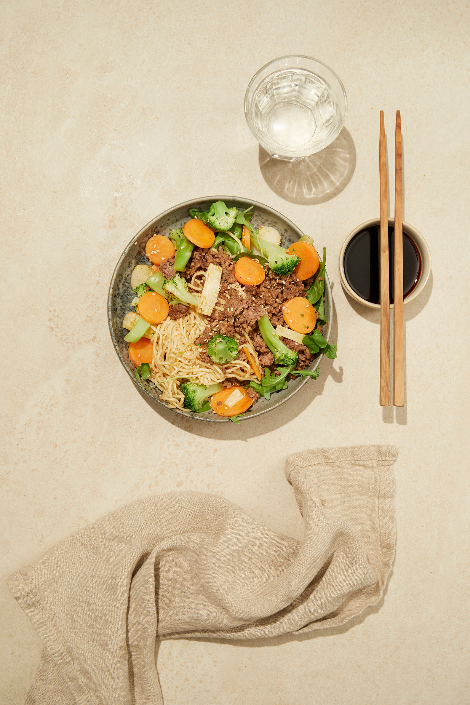 Asiatisk nudelsalat med kjøttdeig, brokkoli, gulrot og sesamfrø. Servert på mørk tallerken, på lys bakgrunn, med spisepinner og et glass vann ved siden av.