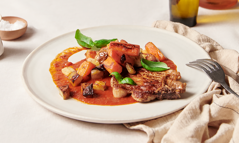Svinekoteletter med ovnsbakte grønnsaker i tomatsaus på en hvit tallerken, med en gaffel på høyre side som ligger på et beige tørkle