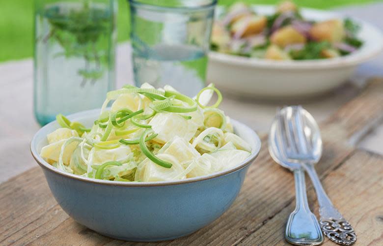 Klassisk kremet potetsalat med grønt eple, toppet med purre. Servert i blå skål, på rustikk trefjøl. Vannglass og serveringsbestikk i bakgrunnen.