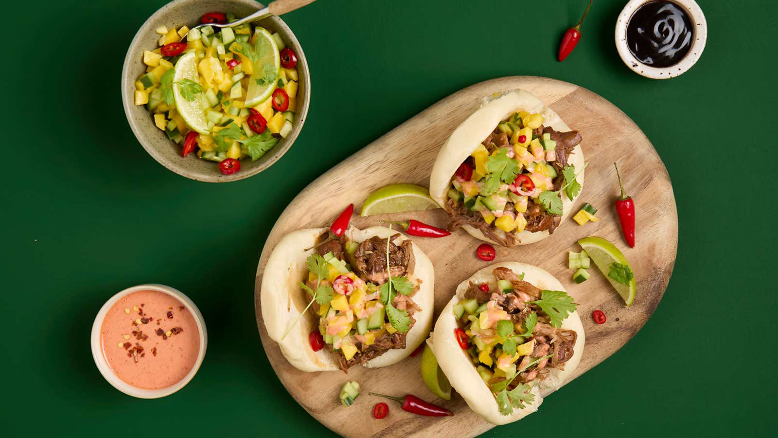Steam buns med pulled pork og mango- og agurksalsa servet på oval trefjøl med limebåter og små røde chili rundt. Skål med salsa på siden.