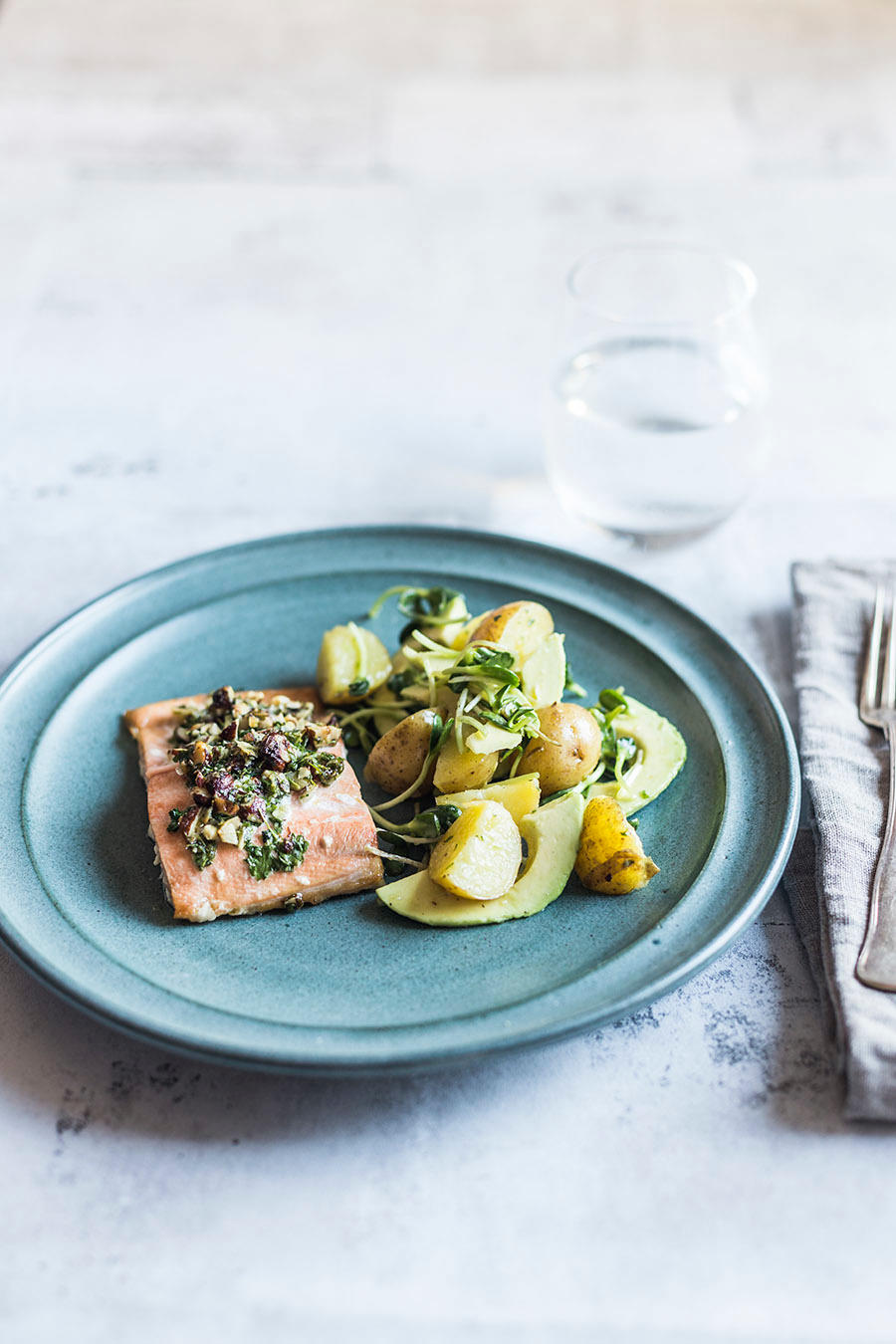 Bakt røye med sitronpoteter, avokado og sprø topping med urter og nøtter, servert på blå asjett på lyst middagsbord med bestikk ved siden av.