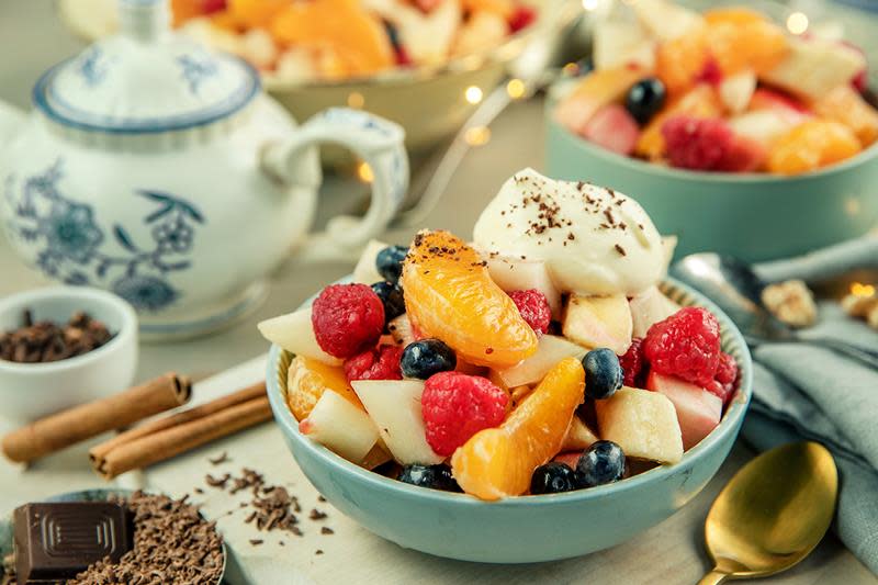 Fruktsalat med julekrydder, hjemmelaget råkrem og dryss av sjokolade, servert i lyseblå skål på dekket bord med tekanne. Stor serveringsbolle med fruktsalat, og julelys i bakgrunnen.
