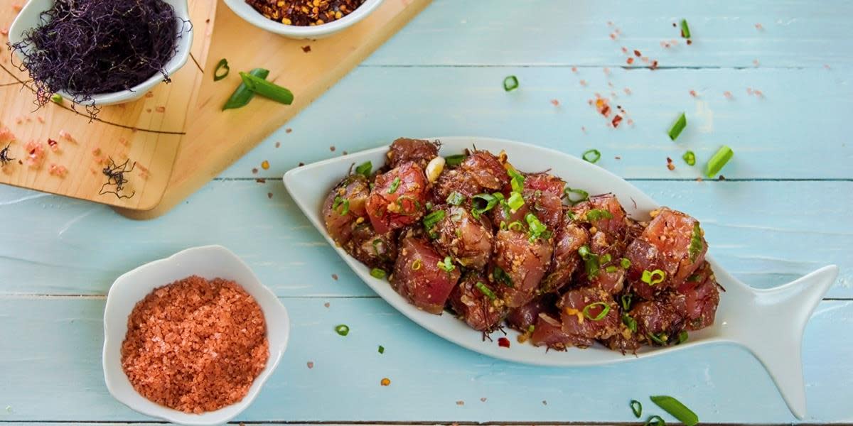 Hjemmelaget poke med laks og rogn servert på fiskeformet hvitt fat, på lyseblått trebord. Finsnittet vårløk strødd over fisken, og små skåler med tilbehør rundt.