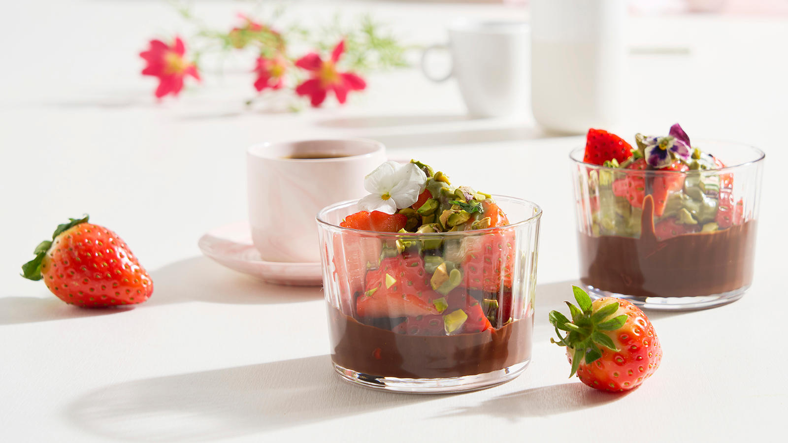 Jordbærdessert med pistasjkrem og sjokolade servert i lite dessertglass med hakkede pistasjnøtter og spiselige blomster. Rosa kopp i bakgrunnen.