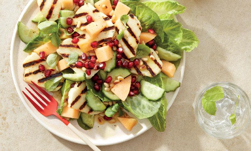 Grillet halloumisalat med melon; grønn salat med grillet halloumi, agurk, granateplefrø og cantaloupemelon på hvit middagstallerken. Lys bakgrunn.