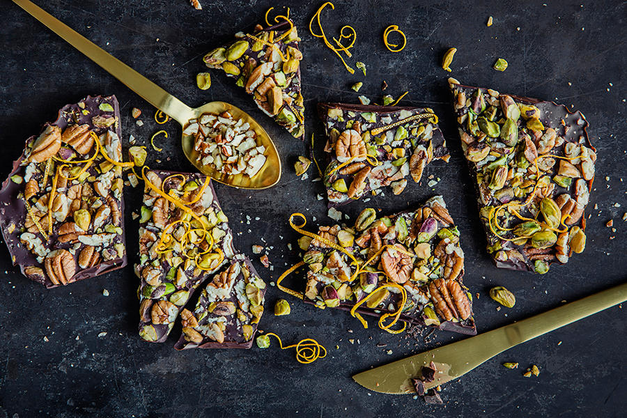 Sjokoladebark med pistasjenøtter, appelsinskall og pekannøtter delt i biter og servert på svart bakgrunn med gullbestikk. Revet appelsinskall drysset over.