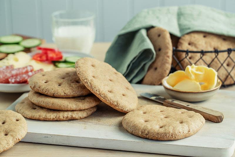 Grove hjemmelagede polarbrød på fjøl, og i kurv, med grønt kjøkkenhåndkle over. Grove polarbrød med pålegg, og et melkeglass i bakgrunnen.