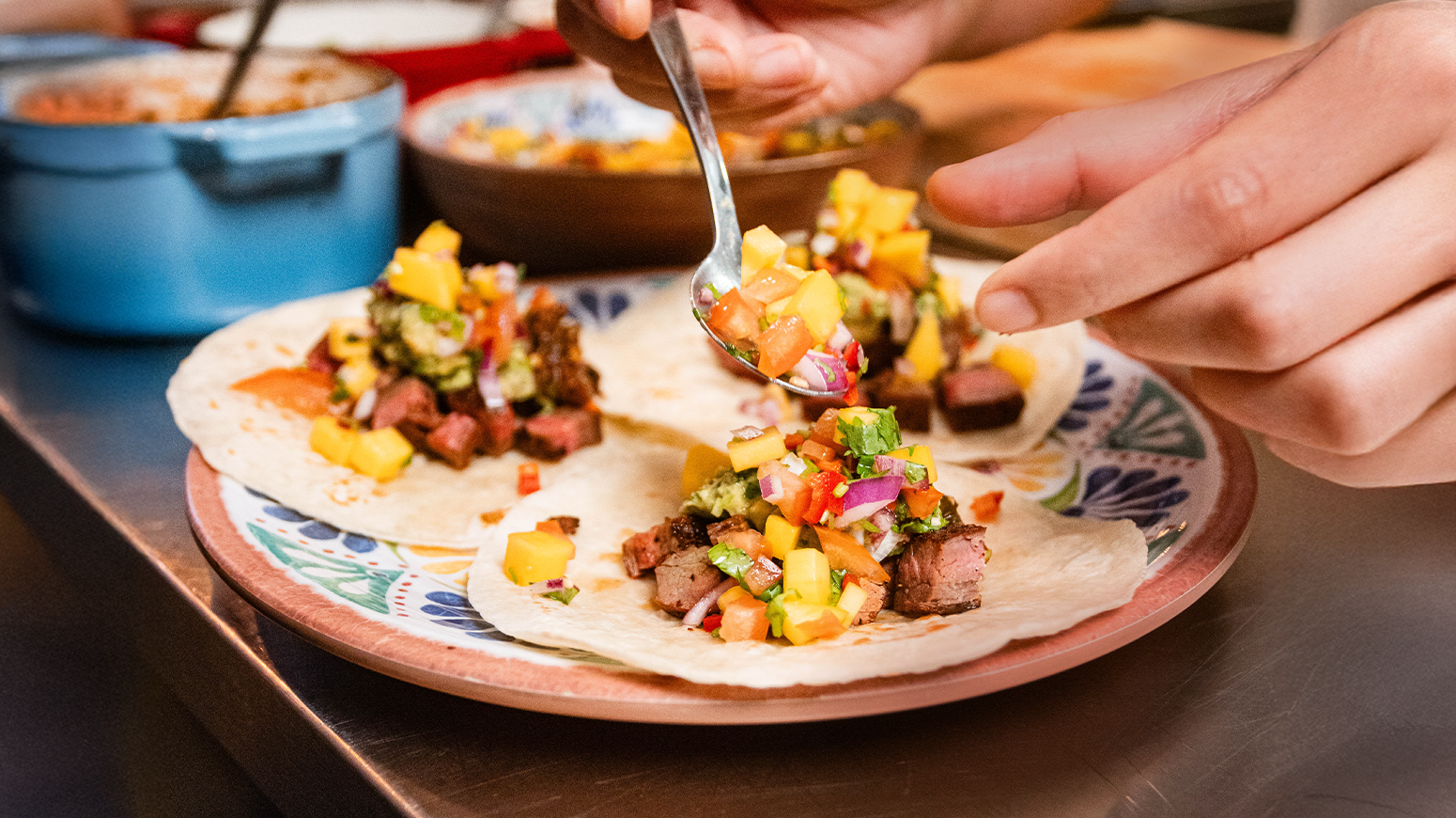 Sommertaco med grillet flank steak, salsa roja og mango pico de gallo servert på tre små tortillas på fargerikt mønstret keramikkfat.