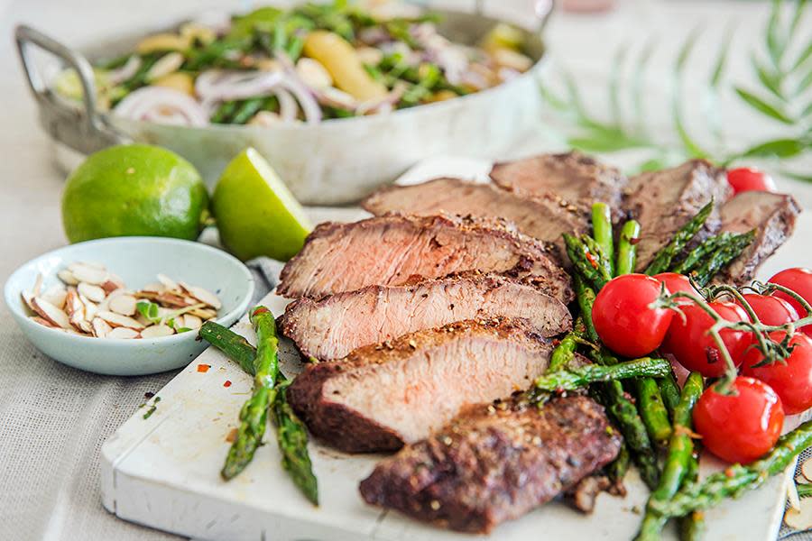 Flat iron steak servert på hvit trefjøl med grillet asparges og cherrytomater. Form med potetsalat med aspargesbønner og mandler i bakgrunnen.