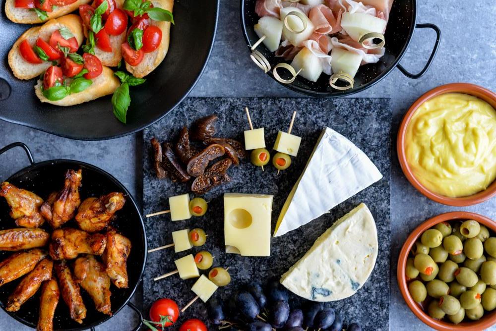 Oversiktsbilde av tapasretter i serveringsskåler på bord. Bruschetta, melon med spekeskinke, hjemmelaget aioli, kyllingklubber ostefat og oliven.