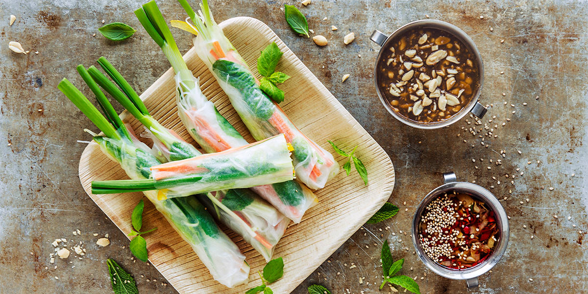Fat med ferske vårruller fylt med grønnsaker og urter. Ved siden av står en skål med peanøttsaus og chilisaus.