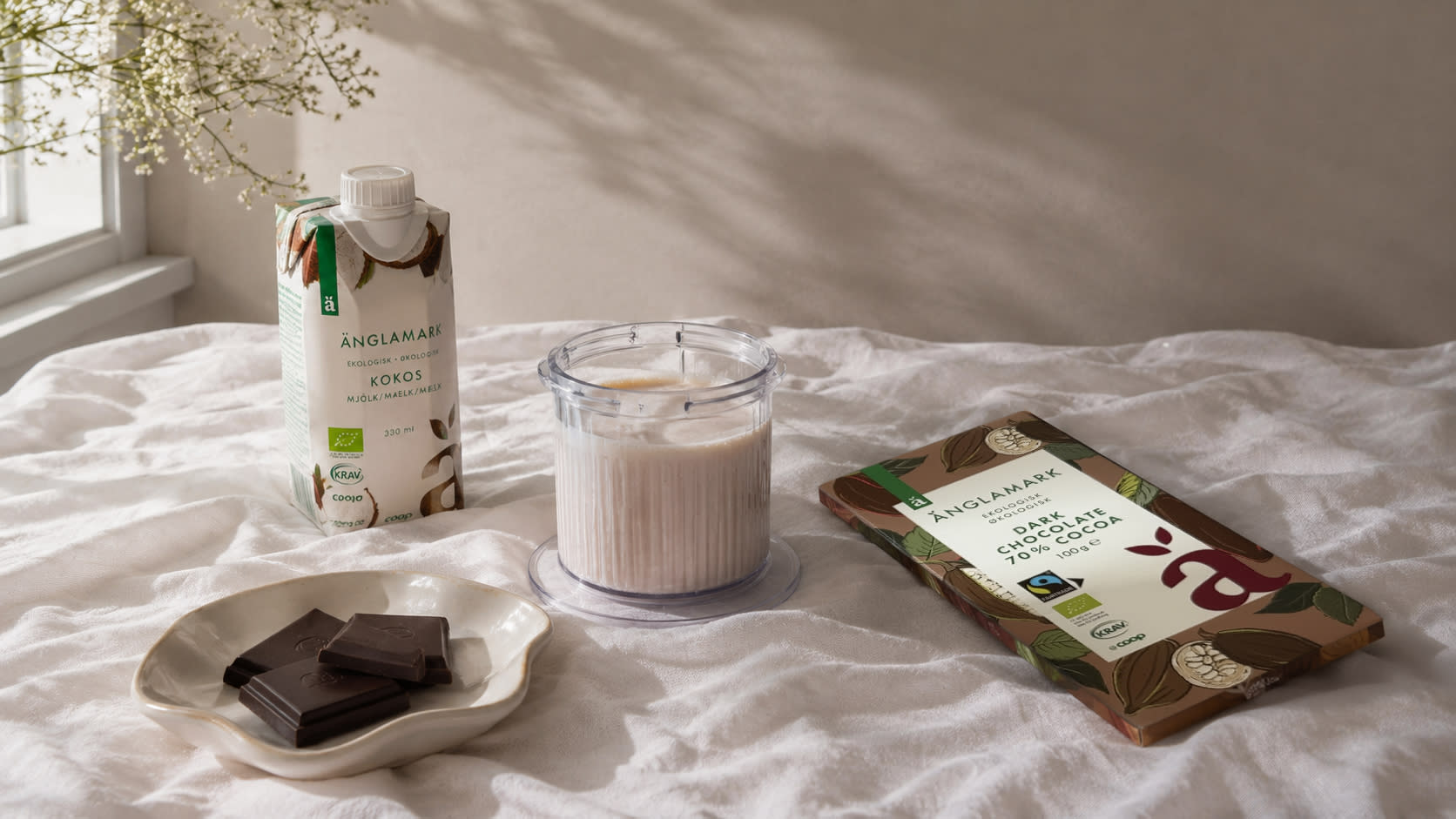 Banan- og kokosis i glass med Änglamark kokosmelk i bakgrunnen og Änglamark Dark chocolate 70% på siden.