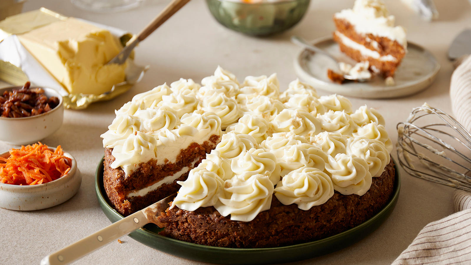 Krydret gulrotkake med ostekrem servert på mørkegrønn tallerken. Skål med revet gulrot, pakke med smør og skål med ostekrem i bakgrunnen.