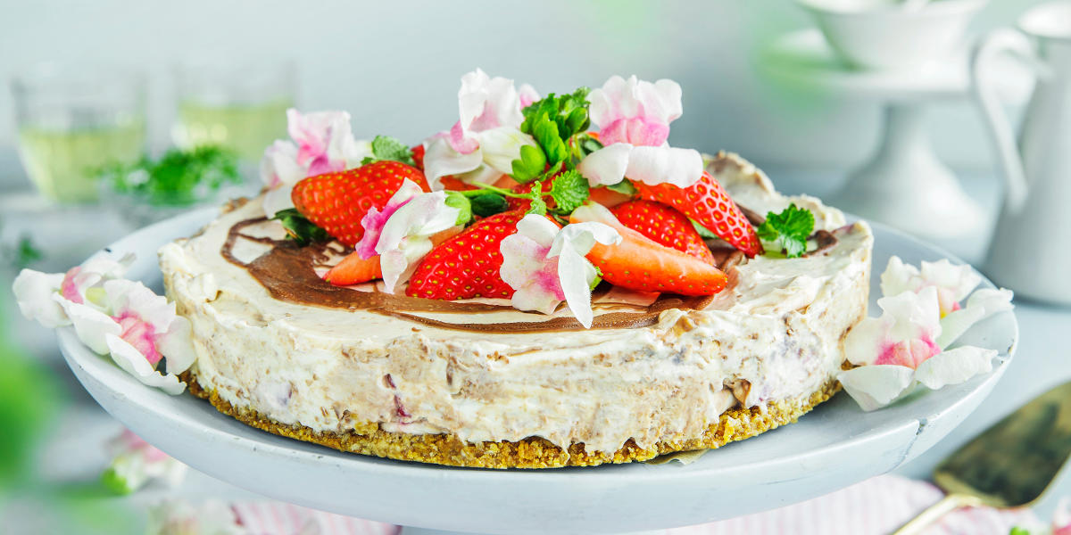 Vidunderlig enkel ostekake med jordbær og melkesjokolade servert på lyst stettfat med spiselige blomster på toppen. Kaken står på dekket kaffebord.