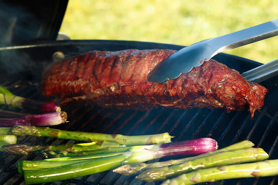 Grillklype som løfter et tykke godt stekt kjøtt fra grillen. Foran steker asparges og vårløk.