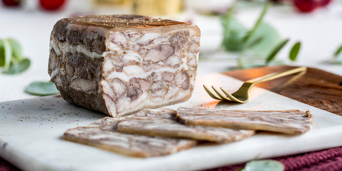 Hjemmelaget sylte med tynnribbe, servert på hvit marmorfjøl med skiver av sylte dandert foran. Gullfarget gaffel ved siden av, og grønt i bakgrunnen.