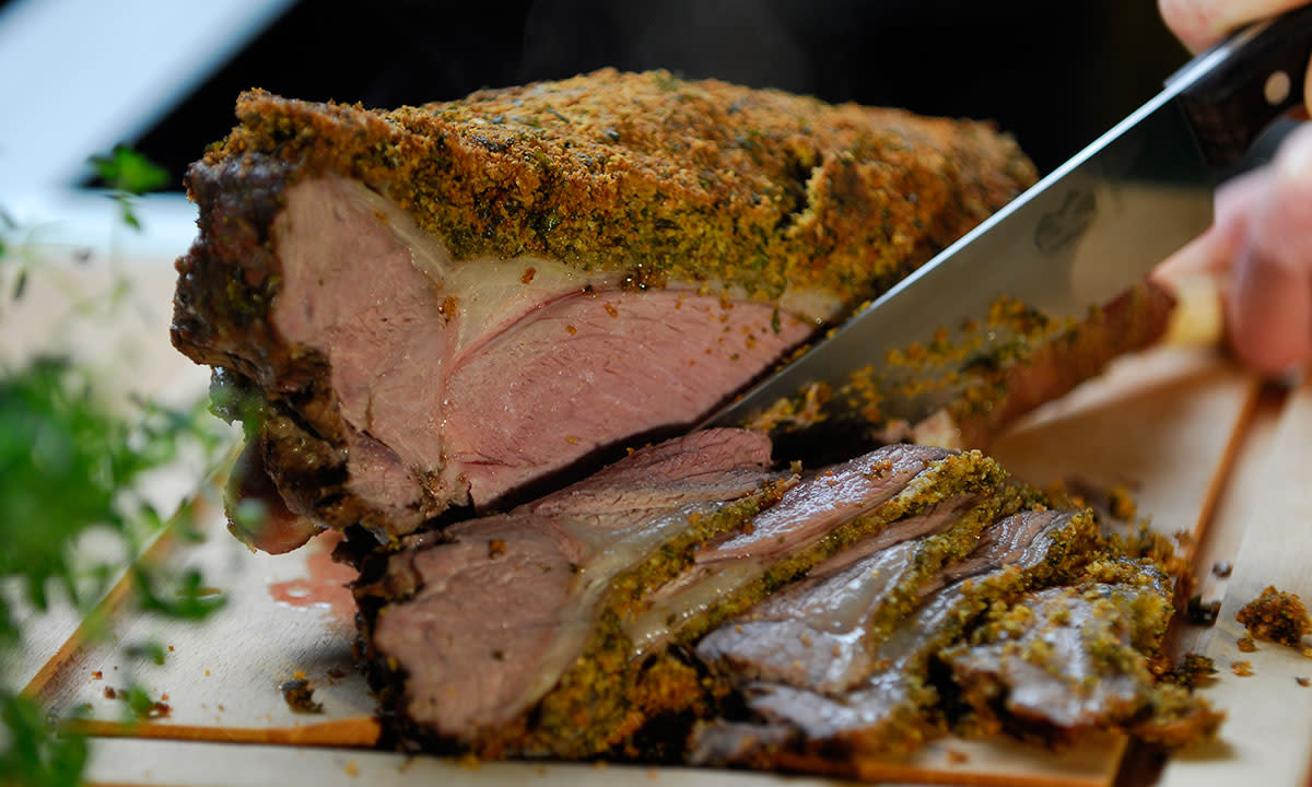 Saftig fransk lammelår med urtelokk skjæres opp på trefjøl. Ferske urter i forkant.