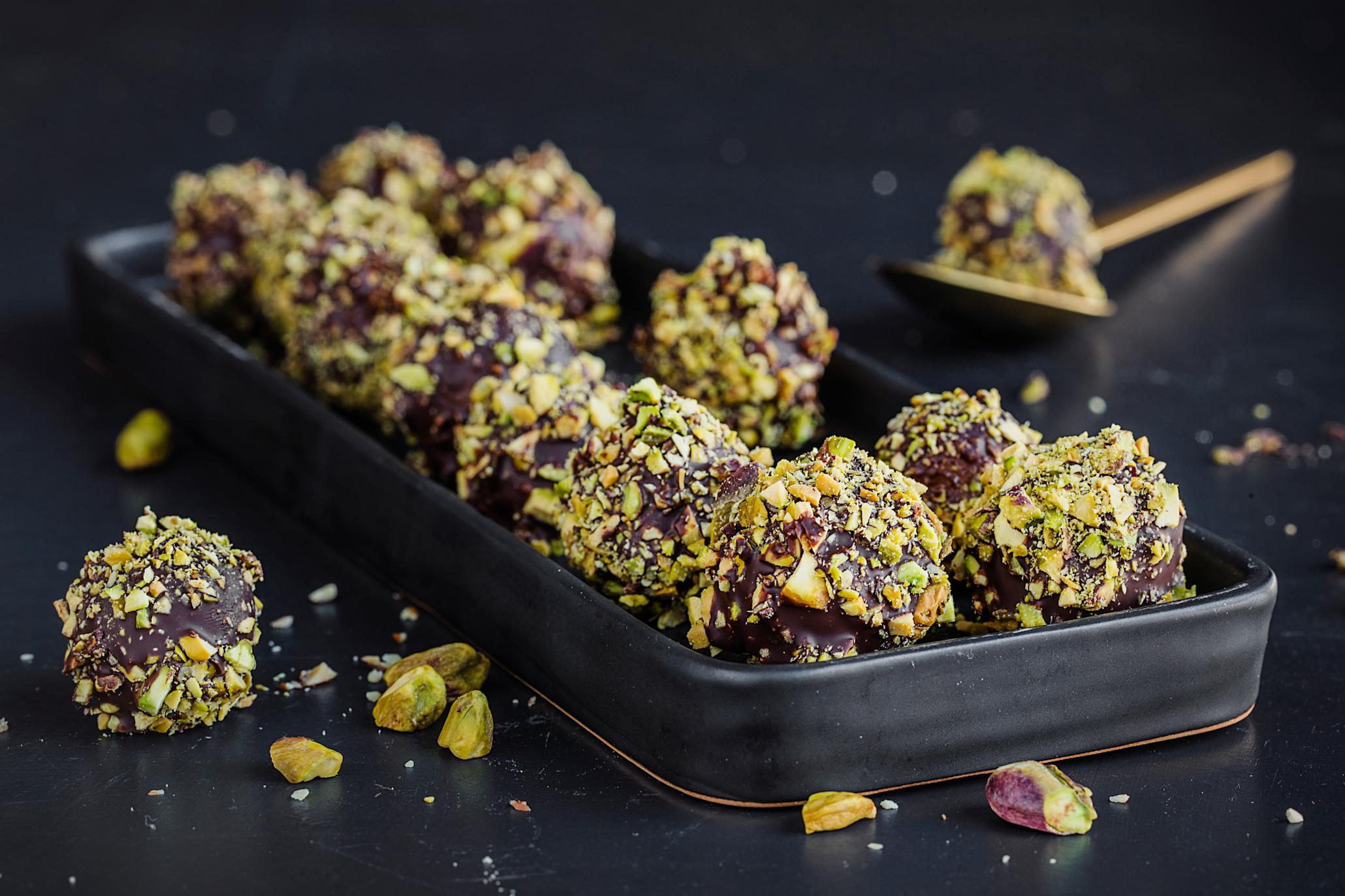 Marsipankuler med sjokolade og pistasje lagt på to rekker på et svart keramikkfat, med grønne pistasjenøtter strødd rundt.