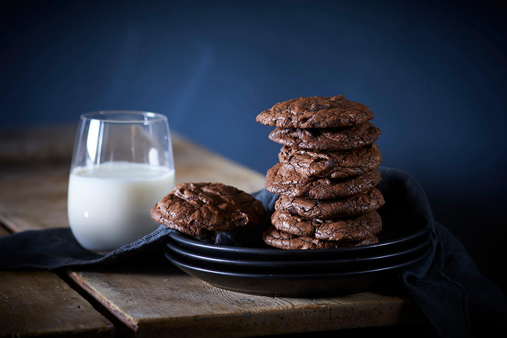 Hjemmelagede browniecookies stablet i tårn på en stabel svarte asjetter, og servert med et glass melk på siden.