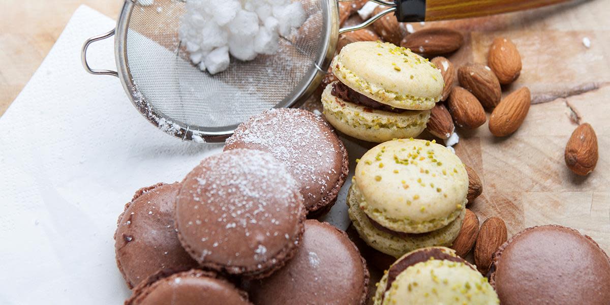 Hjemmelagede makroner i to forskjellige farger servert på hvitt bakepapir på trebord. Liten sikt med melis, og hele mandler med skall ved siden av.