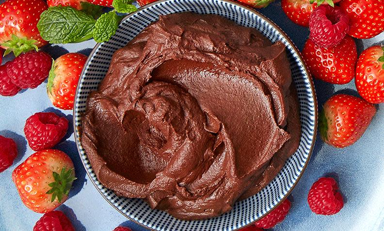 Sjokolademousse laget med avokado, servert i blåstripet skål med ferske jordbær og bringebær rundt. Liten kvast mynte på siden.