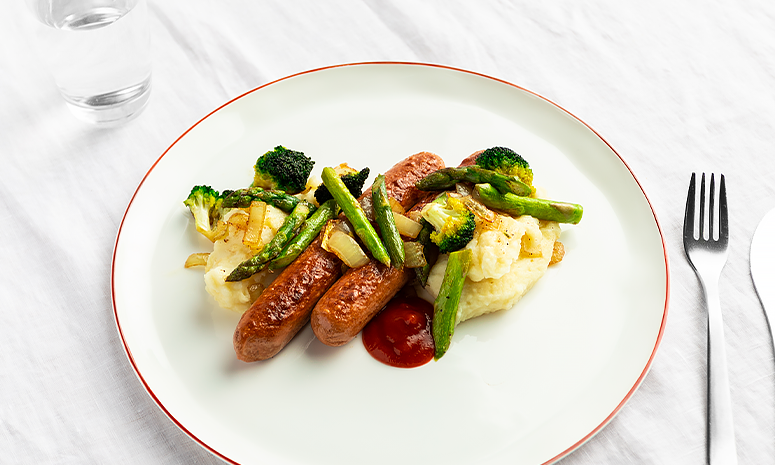 Vegetarpølser, brokkoli, løk, asparges og potetmos på en hvit tallerken med rød kant. Pyntet med litt ketchup.