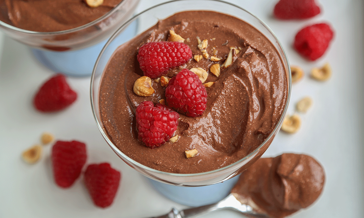 Sjokolademousse med cottage cheese servert i glass med hakkede nøtter og ferske bringebær på toppen. Skje med sjokolademousse, ferske bringebær og hakkede nøtter ved siden av glasset.