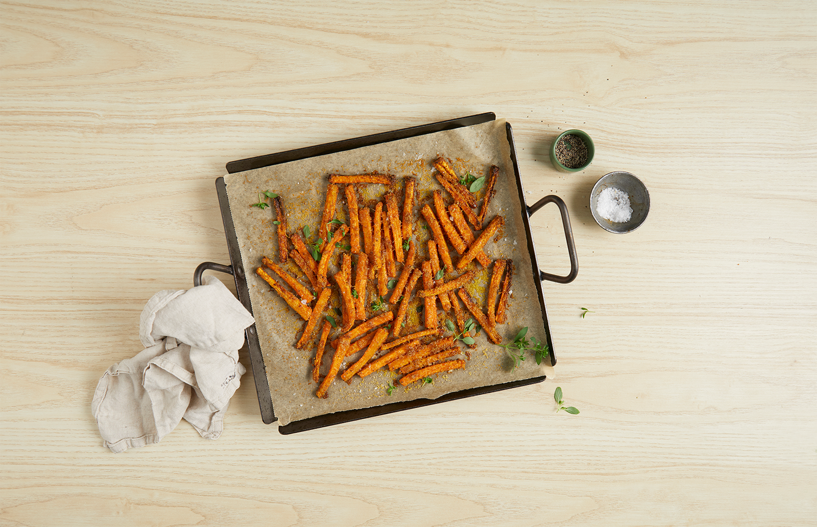 Gulrotfries med parmesan og ferske urter, servert på svart stekebrett med bakepapir. Brettet står på lyst trebord og det er to små skåler med salt og pepper ved siden av.