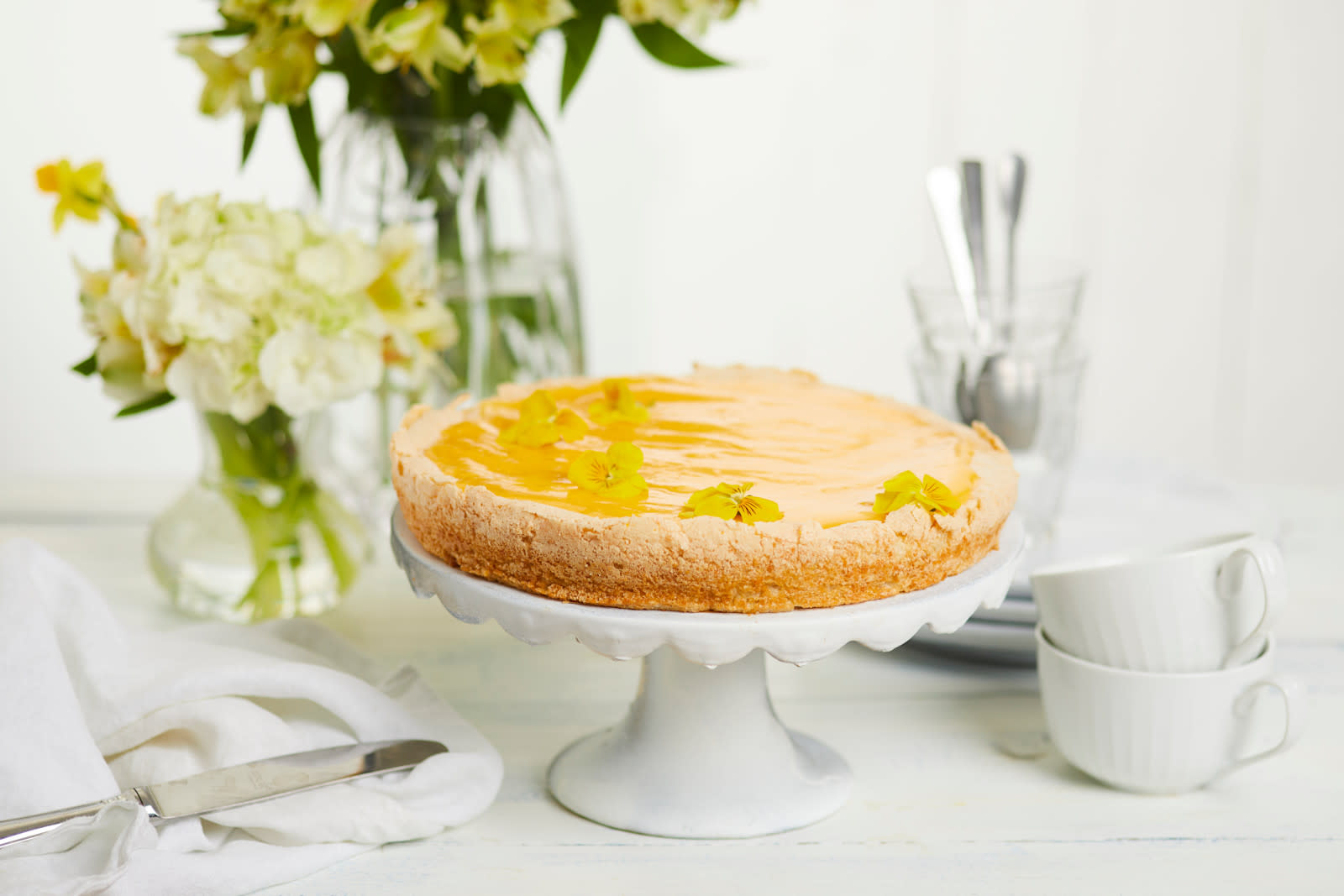 Suksessterte med gul krem pyntet med gule blomster og servert på hvitt stettfat på dekket kaffebord. To vaser lyse blomster i bakgrunnen.