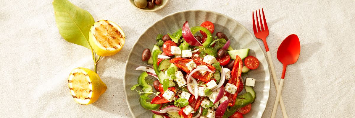 Salat i skaål med paprika, løk, agurk, tomat og fetaost med rødt bestikk og grillet sitron ved siden av
