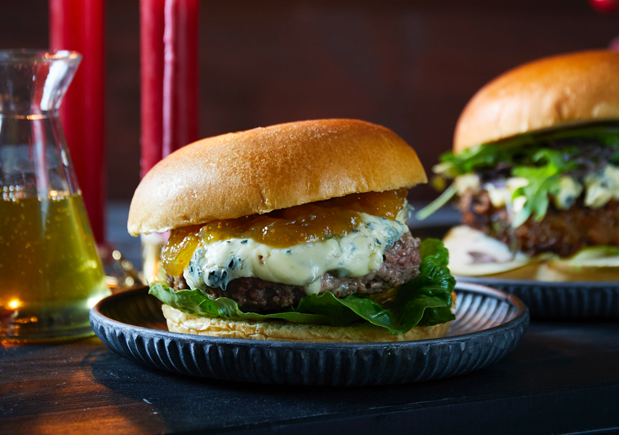  Burger med hjemmelaget fikenmarmelade og smeltet blåmuggost, servert med salatblader på mørk asjett. Asjetten står på mørkt trebord ved siden av en vegetarburger.