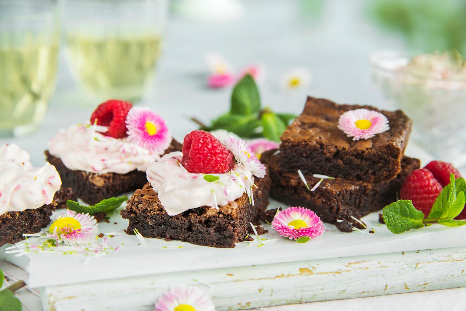 Brownies med bringebærkrem servert i stykker på hvit trefjøl med friske bringebær, mynteblader og blomster. To glass med drikke i bakgrunnen.