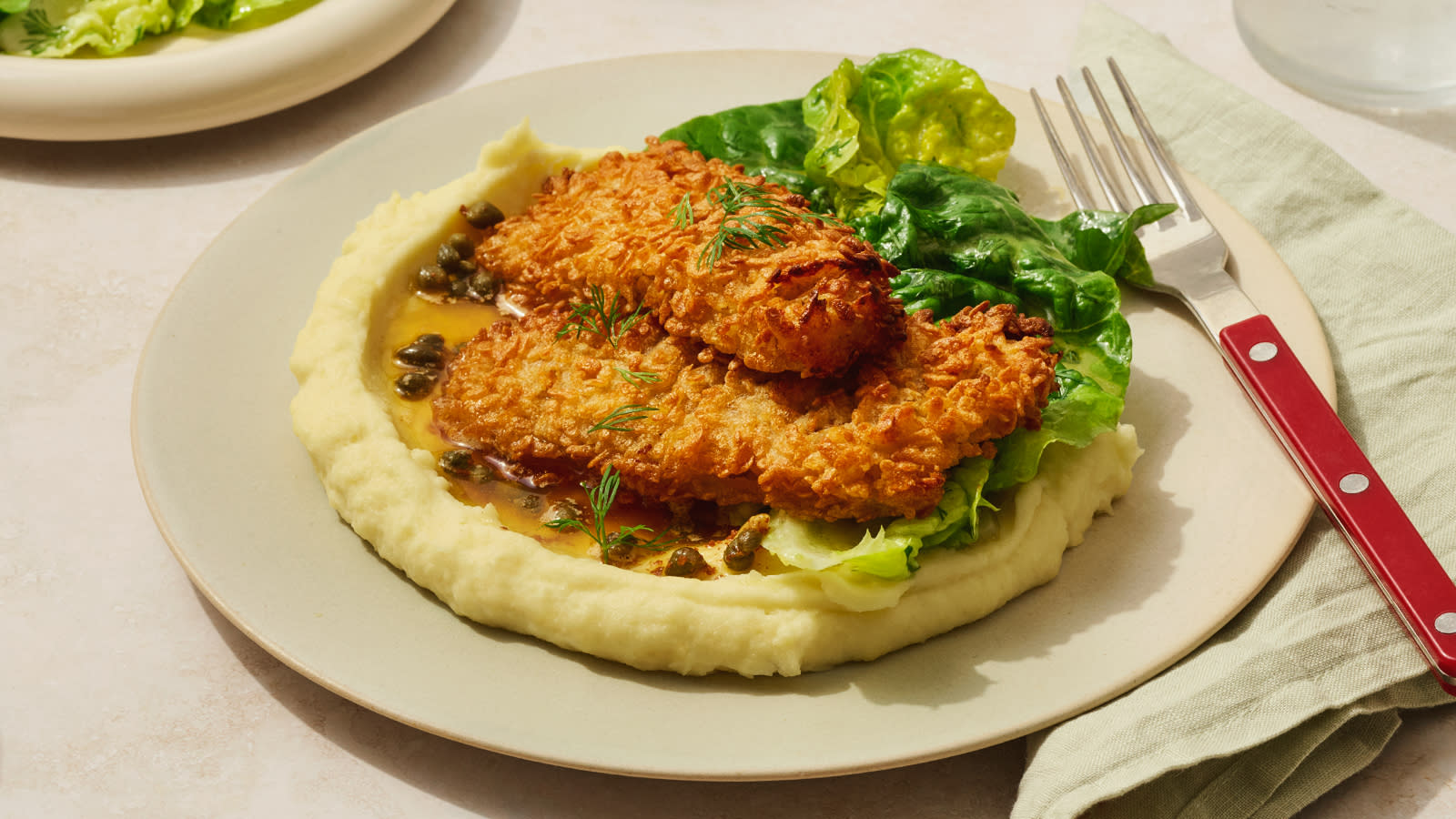 Sprøbakt torsk med dillvinaigrette og potetstappe servert med hjertesalat og kapers på beige tallerken med rød gaffel ved siden av.