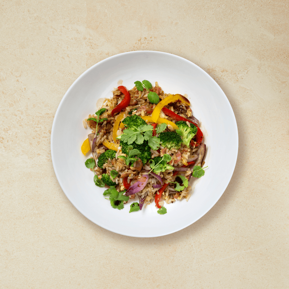 Stekt ris med bacon, brokkoli, rød paprika, gul paprika og rødløk servert i hvit skål med koriander på toppen.