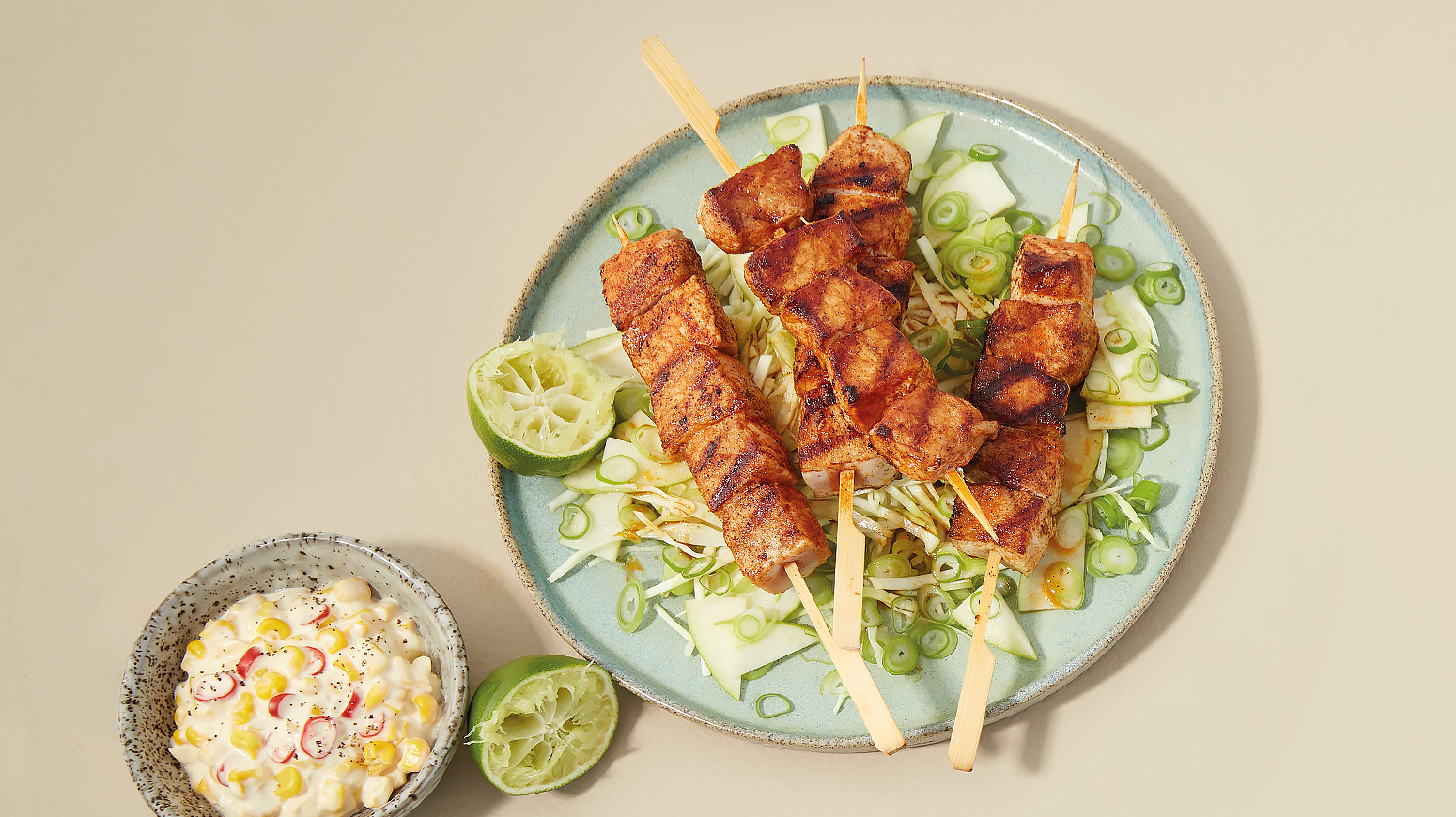 Grillspyd med svinekjøtt og grønn råkost servert på lys turkis tallerken med to halve lime. Liten skål med maisrøre ved siden av.