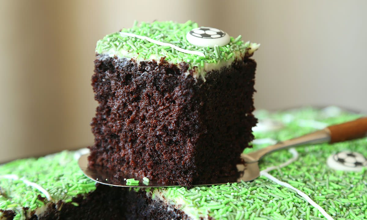 Ekstra saftig sjokoladekake i langpanne pyntet med Dr.Oetker fotballpynt med grønt kakestrø-gress, fotballer og opptegning av fotballbane. Ett stykke av kaken er skåret ut og løftes opp med kakespade.