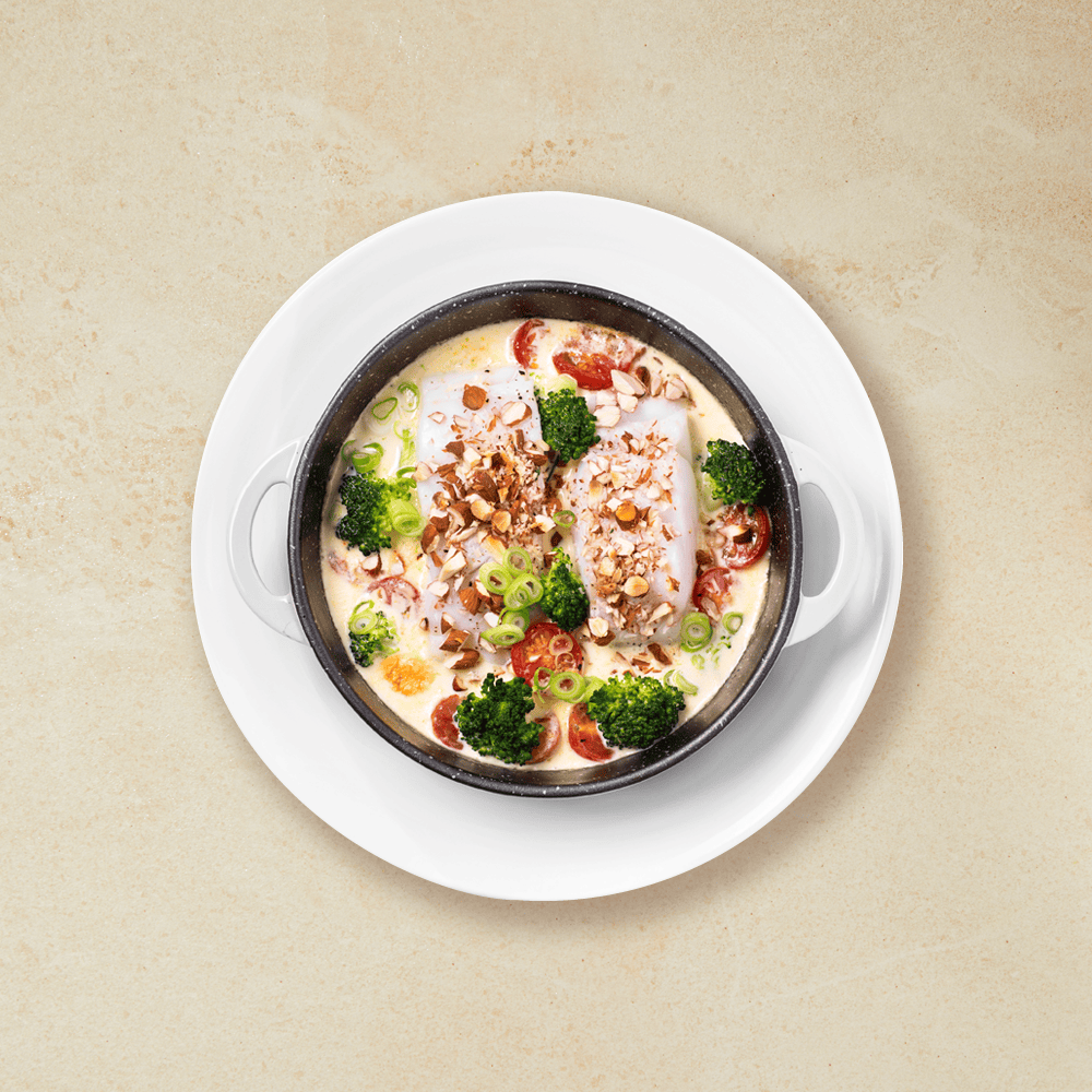 Fløtebakt torsk i form med brokkoli og tomater, med mandler strødd over, servert på hvit tallerken. Beige bakgrunn.