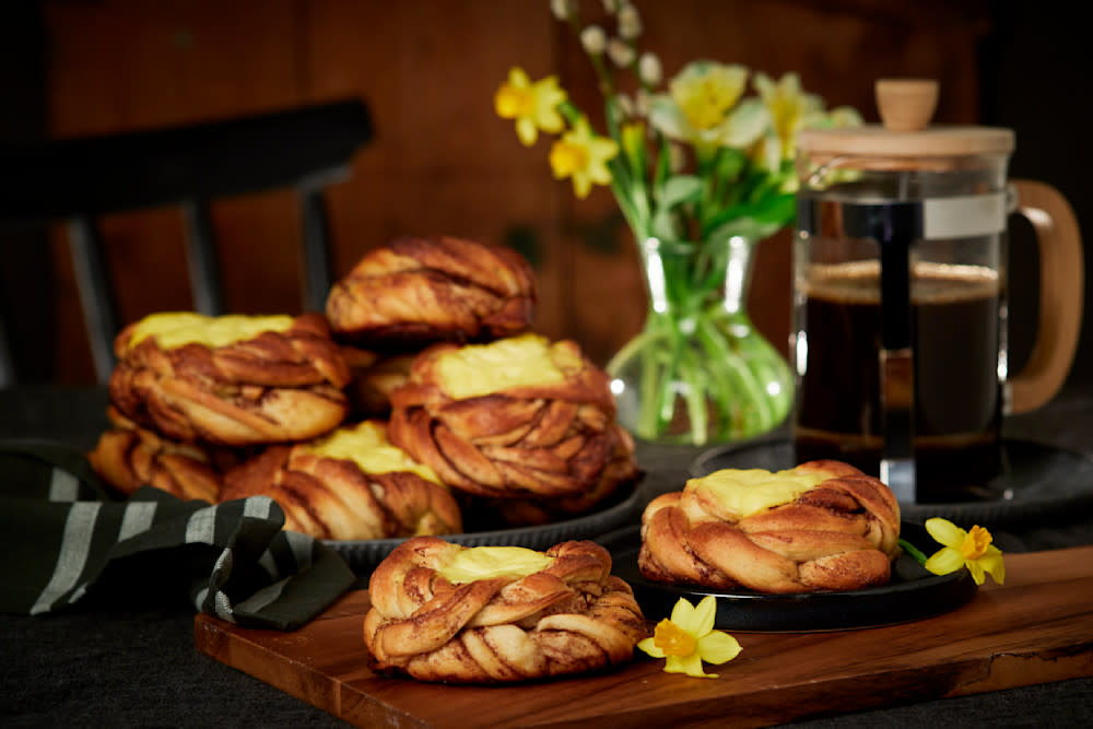 Kanelboller med vaniljekrem servert på både mørk tallerken og mørk trefjøl. Presskanne med kaffe, og vase med påskeliljer i bakgrunnen.