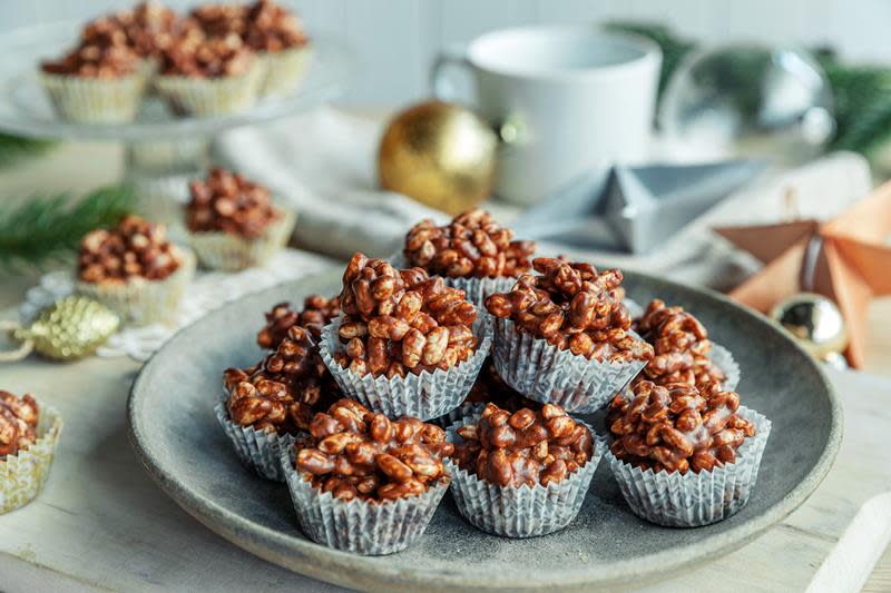 Et lite tårn risboller med sjokolade servert på et grått fat. Omringet av julepynt, julekuler og granbar.