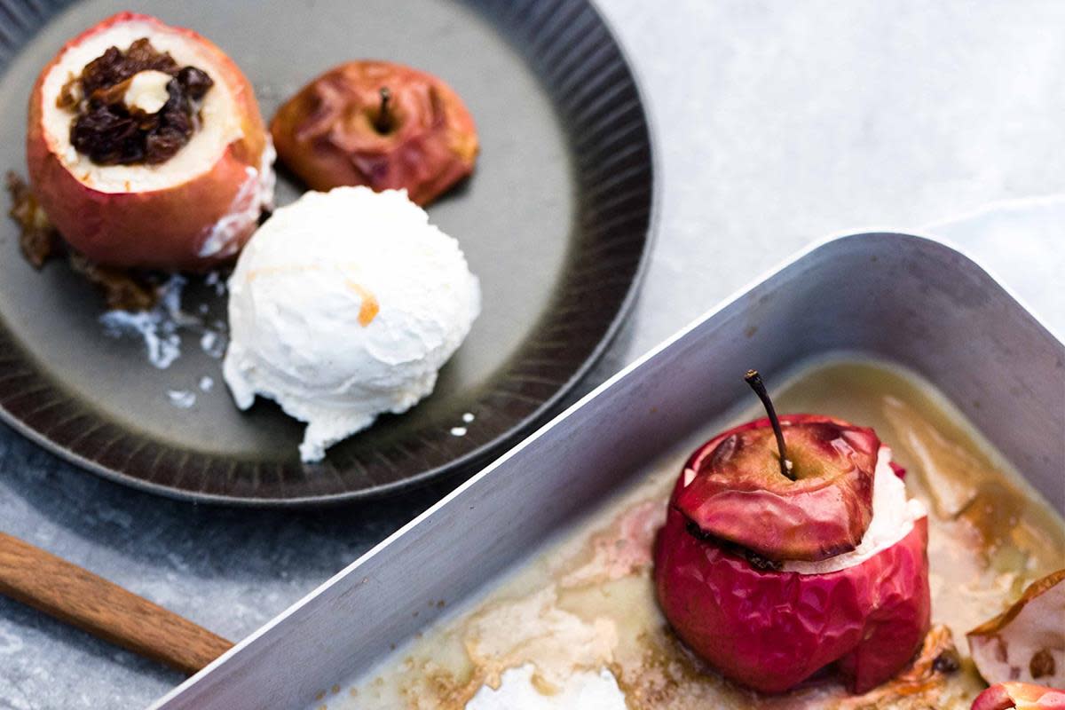 Ovnsbakte epler med rosiner og mandler, servert på mørk tallerken med en kule vaniljeis. Ildfast form med ovnsbakt eple i forgrunnen.