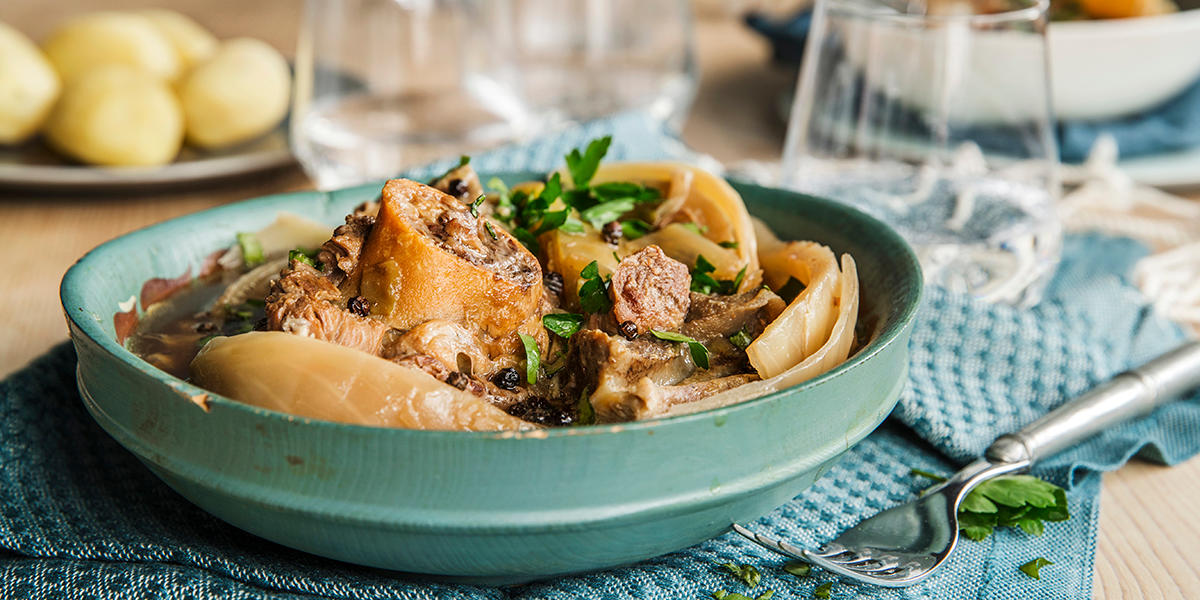 Smaksrik fårikål med lam, kål, hel pepper og bladpersille servert i turkis treskål. Gaffel på siden og poteter i bakgrunnen.