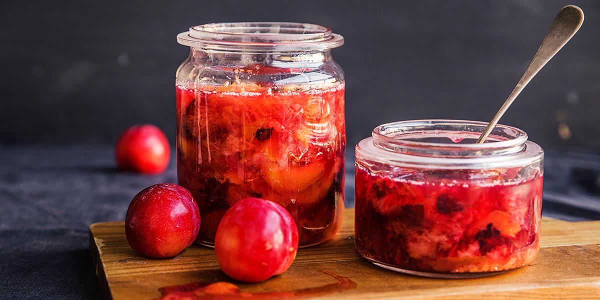 To glass med rødlig plommesyltetøy, servert på mørk trefjøl. Friske plommer ved siden av. Mørk bakgrunn.