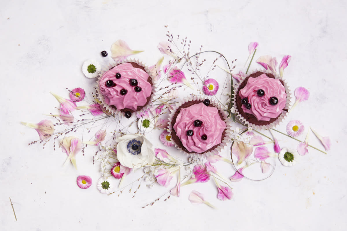 Sticky sjokolademuffins med solbærkrem pyntet med solbær og servert på et underlag av rosa og hvite blomster.