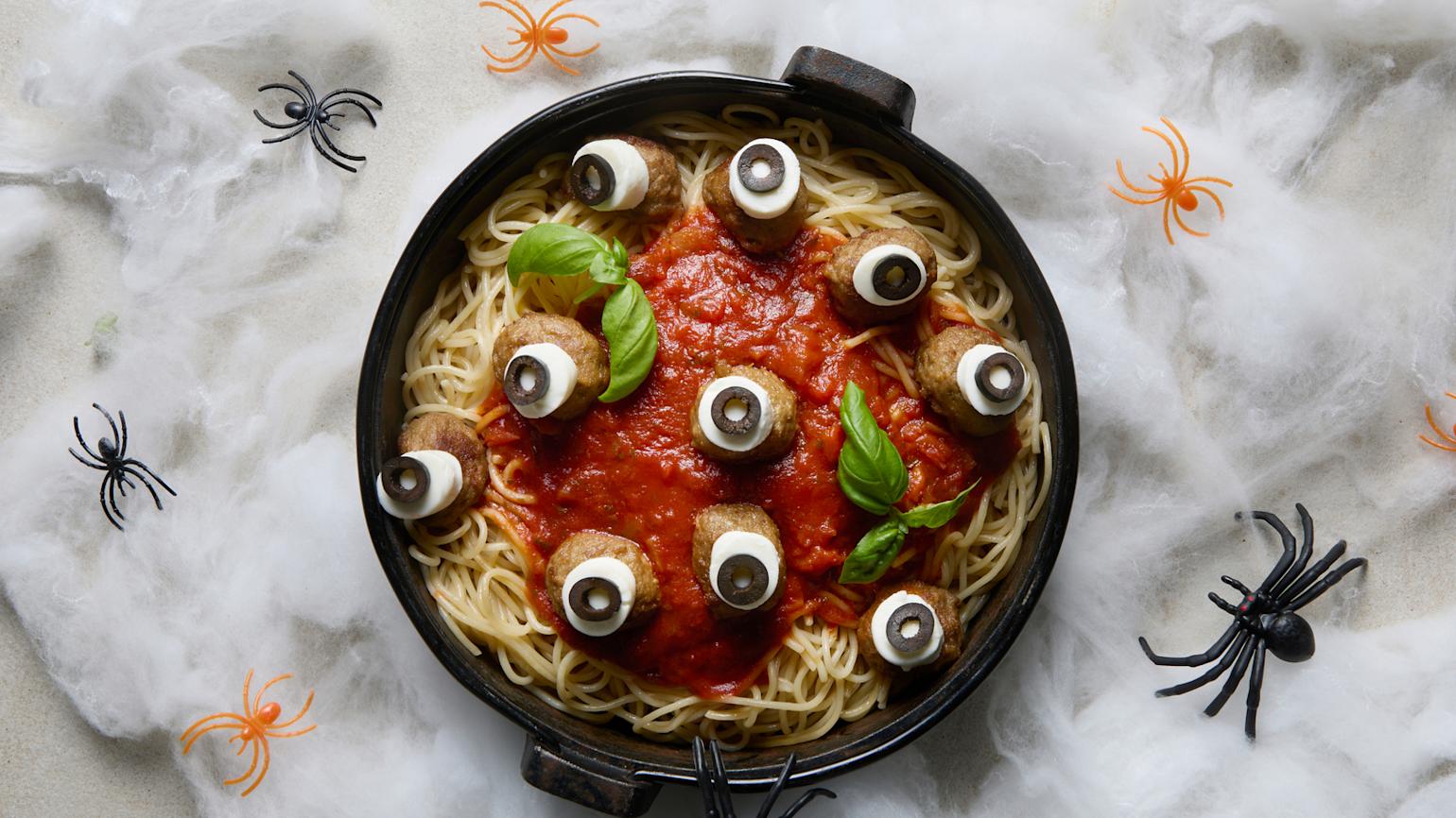 Skummel Halloween spaghetti servert i svart kjele med kjøttboller, mozzarella, pastasaus og frisk basilikum. Spindelvev og plastederkopper i bakgrunnen.